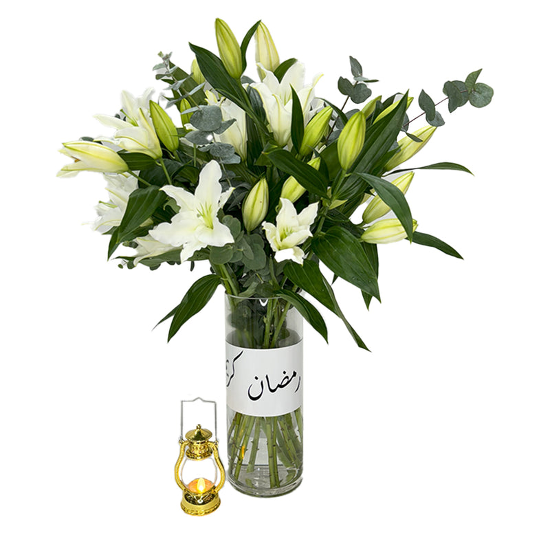 Fresh white lilies in a glass vase with lush greenery and a decorative lantern near by.