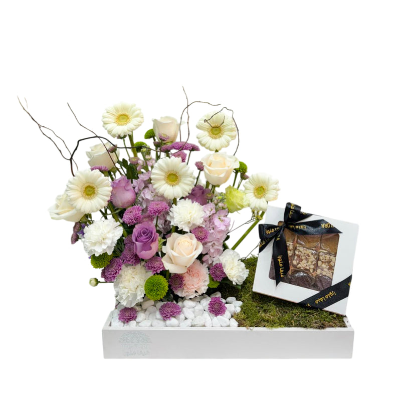 Pastel gerberas, roses, hydrangeas, and carnations arranged atop white stones and moss in a Viva Flora box, with a tied chocolate box.