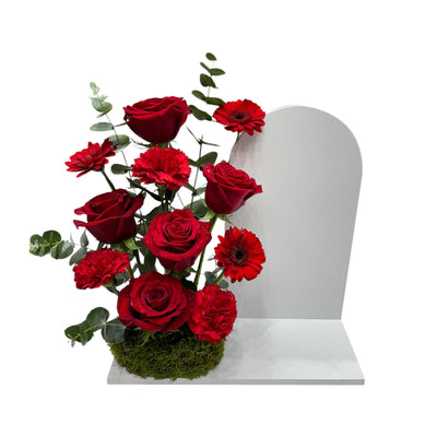 An arrangement of  deep red roses, carnations, and greenery with a white board on the right.