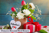 A bouquet of red tulips and white roses in a vase with a Valentine's Day heart and a gift box.
