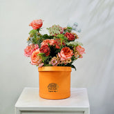 A vibrant orange cylindrical flower box from Viva Flor Tropical, filled with a lush bouquet of pink, coral, and red roses, green foliage, and small white flowers, displayed on a white wooden table against a light gray backdrop.