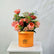 A vibrant orange cylindrical flower box from Viva Flor Tropical, filled with a lush bouquet of pink, coral, and red roses, green foliage, and small white flowers, displayed on a white wooden table against a light gray backdrop.