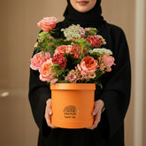 A vibrant orange cylindrical flower box from Viva Flor Tropical, filled with a lush bouquet of pink, coral, and red roses, green foliage, and small white flowers, displayed on a white wooden table against a light gray backdrop.