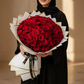 A large round bouquet of deep red roses wrapped in white paper with gold geometric borders, displayed on a round pedestal against a warm beige background.
