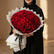 A large round bouquet of deep red roses wrapped in white paper with gold geometric borders, displayed on a round pedestal against a warm beige background.