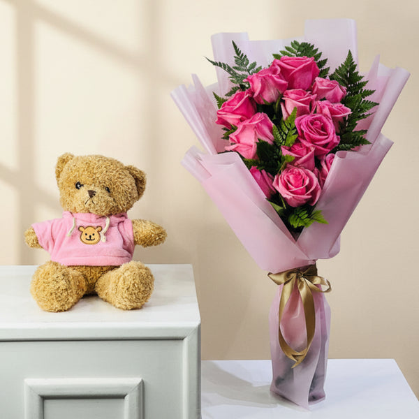 A bouquet of pink roses wrapped in pink paper with a gold ribbon, placed next to a plush teddy bear wearing a pink shirt on white tables against a light gray wall.