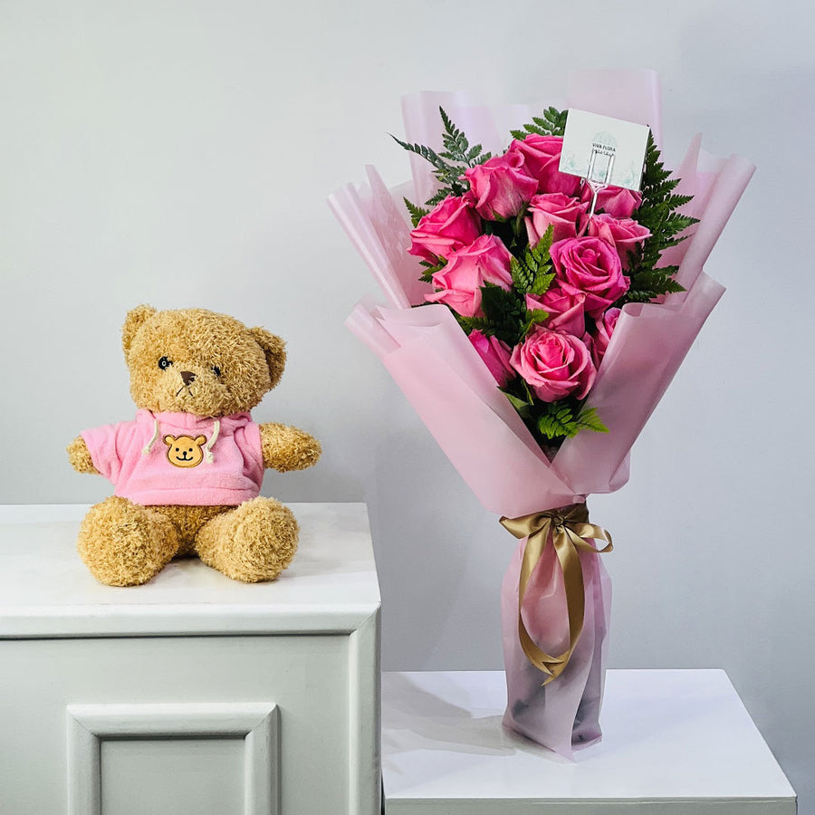 A bouquet of pink roses wrapped in pink paper with a gold ribbon, placed next to a plush teddy bear wearing a pink shirt on white tables against a light gray wall.