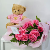 A bouquet of pink roses wrapped in light pink paper with green fern leaves, placed on a white surface, with a brown teddy bear wearing a pink shirt sitting behind it against a plain light background.