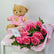 A bouquet of pink roses wrapped in light pink paper with green fern leaves, placed on a white surface, with a brown teddy bear wearing a pink shirt sitting behind it against a plain light background.
