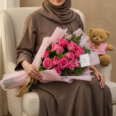 A bouquet of pink roses wrapped in pink paper with a gold ribbon, placed next to a plush teddy bear wearing a pink shirt on white tables against a light gray wall.