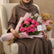 A bouquet of pink roses wrapped in pink paper with a gold ribbon, placed next to a plush teddy bear wearing a pink shirt on white tables against a light gray wall.