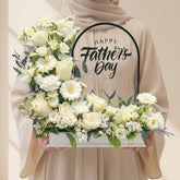 A floral Father's Day cake topper displayed on a white pedestal. The topper features a rectangular white base filled with white roses, daisies, baby's breath, and green foliage, with a black arched sign reading 'Happy Fathers Day'.