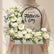 A floral Father's Day cake topper displayed on a white pedestal. The topper features a rectangular white base filled with white roses, daisies, baby's breath, and green foliage, with a black arched sign reading 'Happy Fathers Day'.