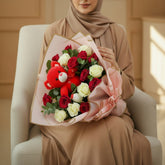 A floral gift arrangement featuring red and cream roses wrapped in pink paper with gold trim, topped with a red teddy bear, displayed on a white pedestal against a plain light gray background.