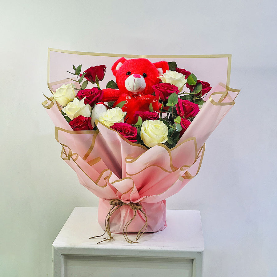 A floral gift arrangement featuring red and cream roses wrapped in pink paper with gold trim, topped with a red teddy bear, displayed on a white pedestal against a plain light gray background.