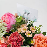 A close-up of a floral arrangement with pink and peach roses, coral carnations, pale daisy-like flowers, green foliage, and a small card reading VIVA FLORA.