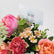 A close-up of a floral arrangement with pink and peach roses, coral carnations, pale daisy-like flowers, green foliage, and a small card reading VIVA FLORA.
