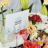 A white printed card with the brand name VIVA FLORA and floral line art, displayed in a clear acrylic stand among a bouquet of flowers including white lilies, pink roses, yellow gerbera daisies, and red carnations.