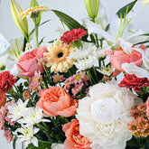 A close-up of a mixed floral arrangement featuring white lilies, peach and pink roses, white and yellow gerbera daisies, red carnations, and green foliage against a light neutral background.