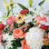 A close-up of a mixed floral arrangement featuring white lilies, peach and pink roses, white and yellow gerbera daisies, red carnations, and green foliage against a light neutral background.