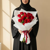 A bouquet of 15 red roses wrapped in white tissue paper and tied with a dark red ribbon, displayed on a white pedestal against a beige wall with soft window shadow patterns.