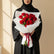 A bouquet of 15 red roses wrapped in white tissue paper and tied with a dark red ribbon, displayed on a white pedestal against a beige wall with soft window shadow patterns.