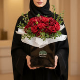 A bouquet of red roses wrapped in white tissue paper and presented in a black square box with the brand name VIVA FLORA, placed on a white cylindrical pedestal against a beige wall with soft window shadows.