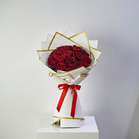 A bouquet of red roses wrapped in white paper with gold trim and a red ribbon, displayed on a white box against a light gray background.