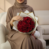 A bouquet of 20 red roses wrapped in white paper with gold trim and tied with a red ribbon, displayed on a white pedestal against a beige background.
