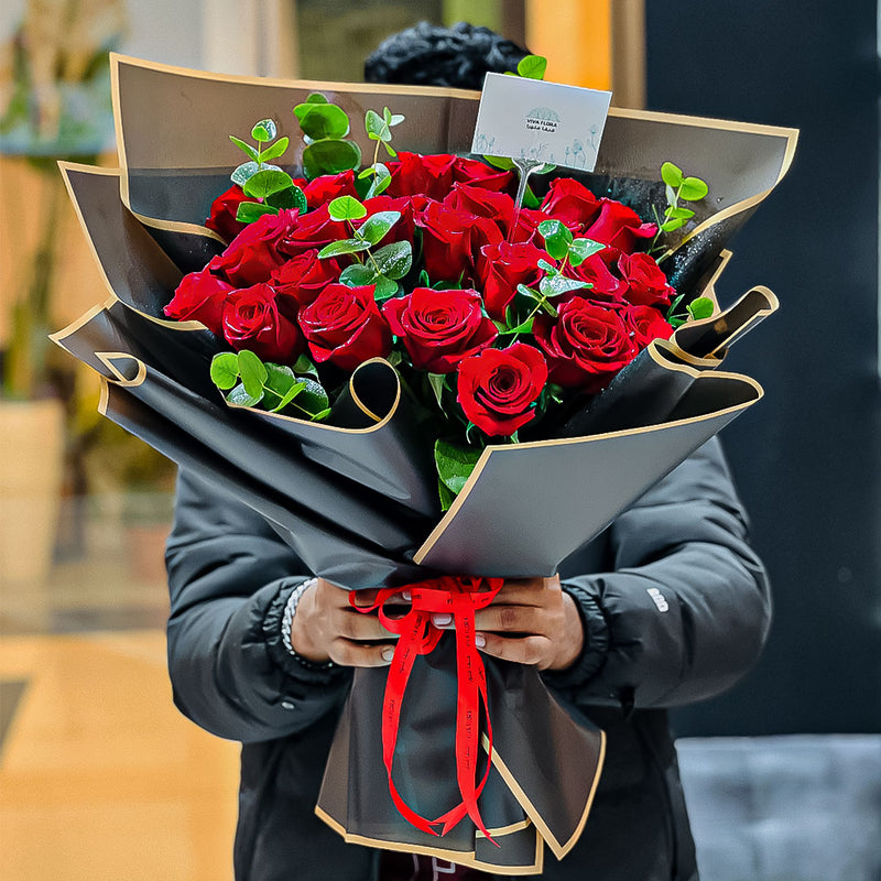 Bouquet of red roses wrapped in black paper with a red ribbon held by a person.