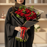 A bouquet of red roses wrapped in black paper with gold trim and tied with a red ribbon, displayed on a white cylindrical pedestal against a warm beige wall with soft window-light shadows.