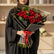 A bouquet of red roses wrapped in black paper with gold trim and tied with a red ribbon, displayed on a white cylindrical pedestal against a warm beige wall with soft window-light shadows.
