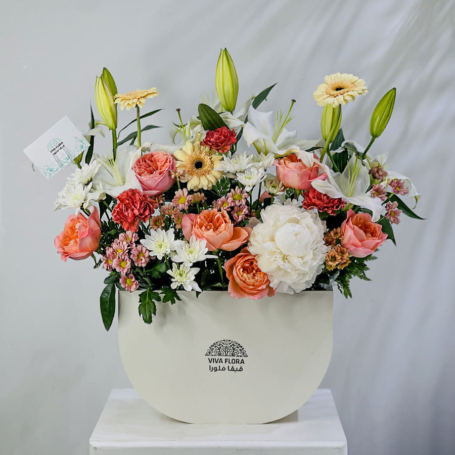 A vibrant floral arrangement of lilies, roses, daisies, and carnations in a white semi-circular planter with the brand name 'VIVA FLORA' printed on the front, displayed on a white pedestal against a light gray backdrop.