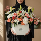 A vibrant floral arrangement of lilies, roses, daisies, and carnations in a white semi-circular planter with the brand name 'VIVA FLORA' printed on the front, displayed on a white pedestal against a light gray backdrop.