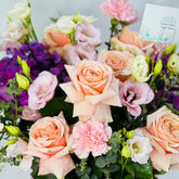 A close-up of a floral bouquet featuring peach and pink roses, light pink carnations, purple lisianthus, white and cream stock flowers, and green buds with eucalyptus leaves. A small card reading Viva Flora is visible at the top right.