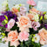 A close-up of a floral bouquet featuring peach and pink roses, light pink carnations, purple lisianthus, white and cream stock flowers, and green buds with eucalyptus leaves. A small card reading Viva Flora is visible at the top right.