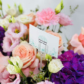 A floral arrangement with pink, peach, purple, and white flowers surrounding a small white card holder printed with the brand name VIVA FLORA.