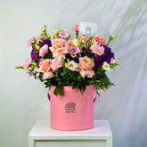A vibrant floral arrangement featuring peach roses, purple lisianthus, pink carnations, and green foliage, presented in a pink cylindrical bucket planter with the brand name 'VIVA FLOR' printed on the front, set on a white pedestal against a light gray background.