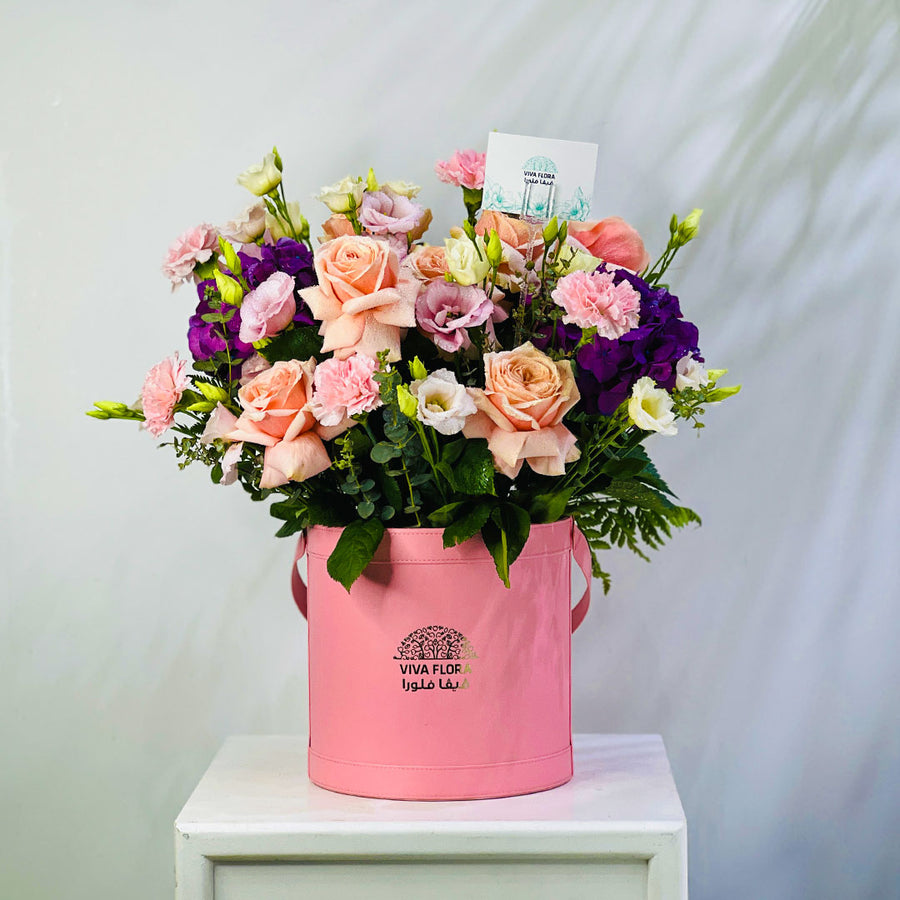 A vibrant floral arrangement featuring peach roses, purple lisianthus, pink carnations, and green foliage, presented in a pink cylindrical bucket planter with the brand name 'VIVA FLOR' printed on the front, set on a white pedestal against a light gray background.