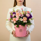A vibrant floral arrangement featuring peach roses, purple lisianthus, pink carnations, and green foliage, presented in a pink cylindrical bucket planter with the brand name 'VIVA FLOR' printed on the front, set on a white pedestal against a light gray background.
