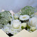 A close-up of a bouquet of white and cream flowers, including roses, daisies, chrysanthemums, and green leafy filler stems, arranged in a cream-colored paper wrap.