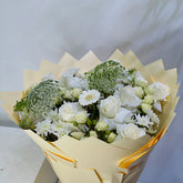 A bouquet of white and pale yellow flowers wrapped in cream-colored paper, featuring white roses, daisies, baby's breath, and green foliage, set against a plain light background.