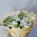 A bouquet of white and pale yellow flowers wrapped in cream-colored paper, featuring white roses, daisies, baby's breath, and green foliage, set against a plain light background.
