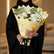 A bouquet of white and light green flowers wrapped in pale yellow paper and tied with a black ribbon, displayed on a white cabinet against a light gray wall.