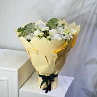 A bouquet of white and light green flowers wrapped in pale yellow paper and tied with a black ribbon, displayed on a white cabinet against a light gray wall.