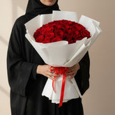 A bouquet of 50 red roses wrapped in white paper and tied with a red ribbon, displayed on a white pedestal against a warm beige background with soft window shadows.