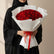 A bouquet of 50 red roses wrapped in white paper and tied with a red ribbon, displayed on a white pedestal against a warm beige background with soft window shadows.