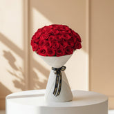 A person wearing a black abaya and hijab holds a large bouquet of red roses wrapped in white paper with a black ribbon featuring gold text, set against a warm beige background.