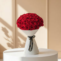 A person wearing a black abaya and hijab holds a large bouquet of red roses wrapped in white paper with a black ribbon featuring gold text, set against a warm beige background.