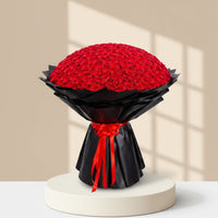 A large bouquet of red roses wrapped in black paper with a red ribbon bow, photographed on a clean white background.